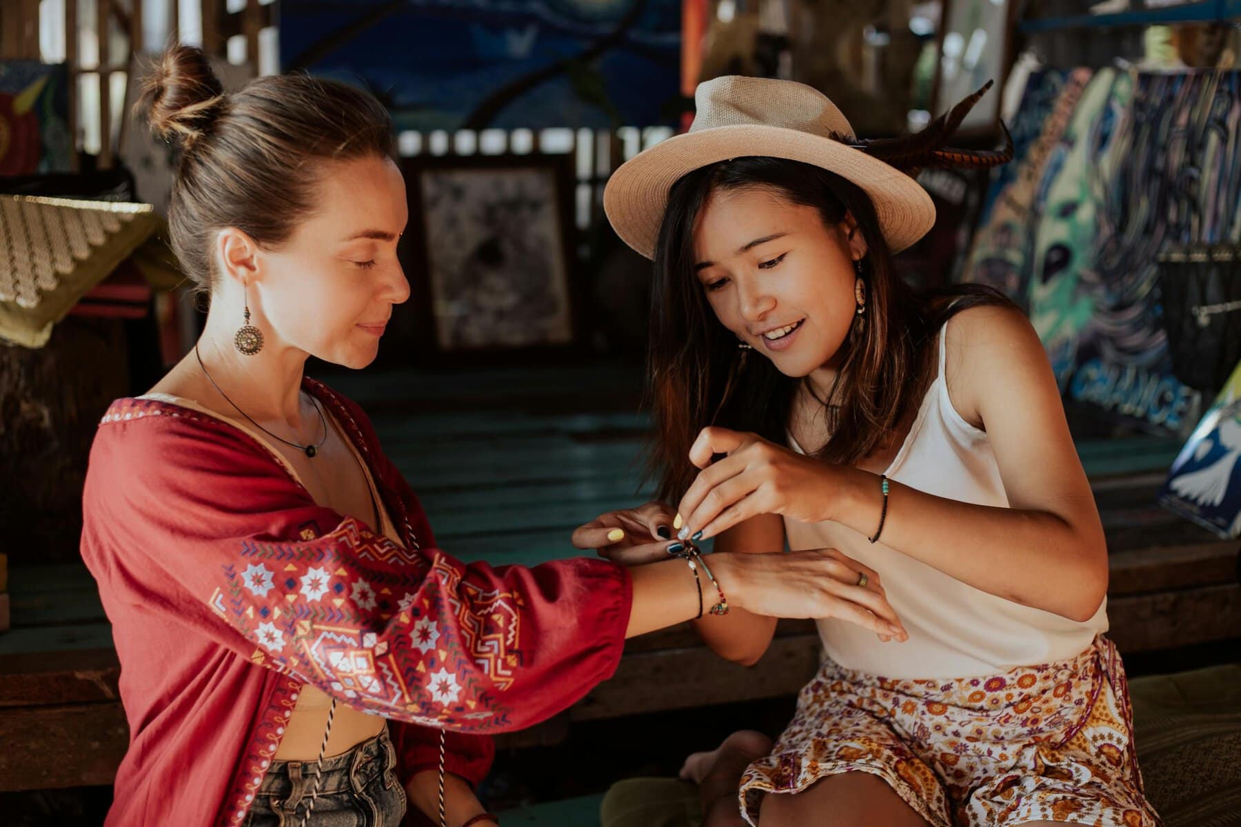 Two women bonding by exchanging friendship bracelets inside a cozy art space.