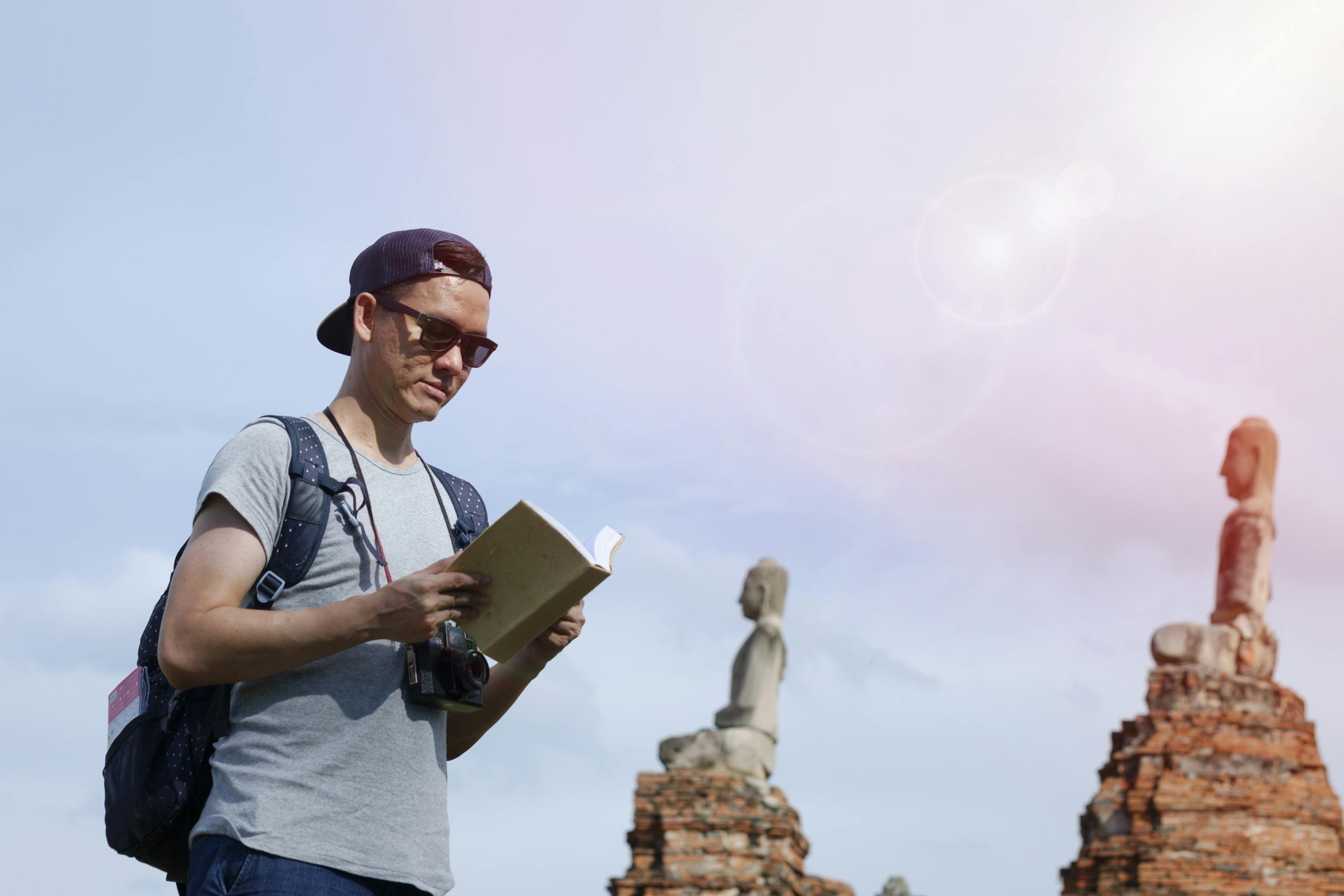 A tourist with sunglasses reads a book near ancient statues under daylight.