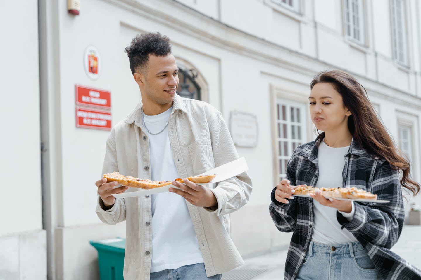 Two young adults enjoying pizza while walking outdoors in the city, creating a joyful street food moment.
