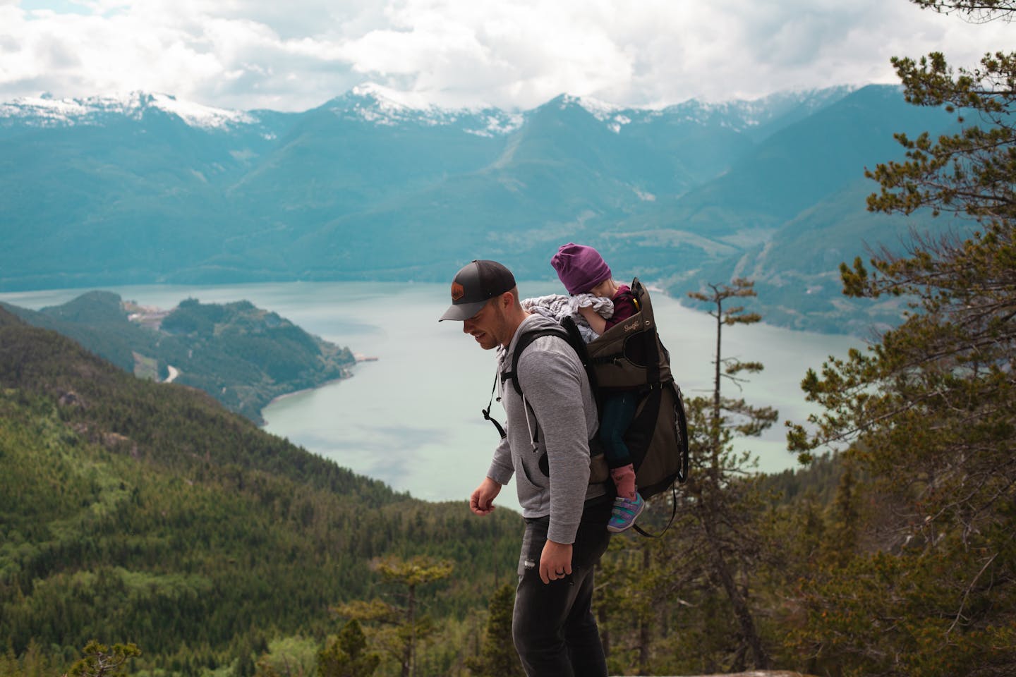 A father carries his child on a mountain hike with breathtaking views of a lake and mountains.