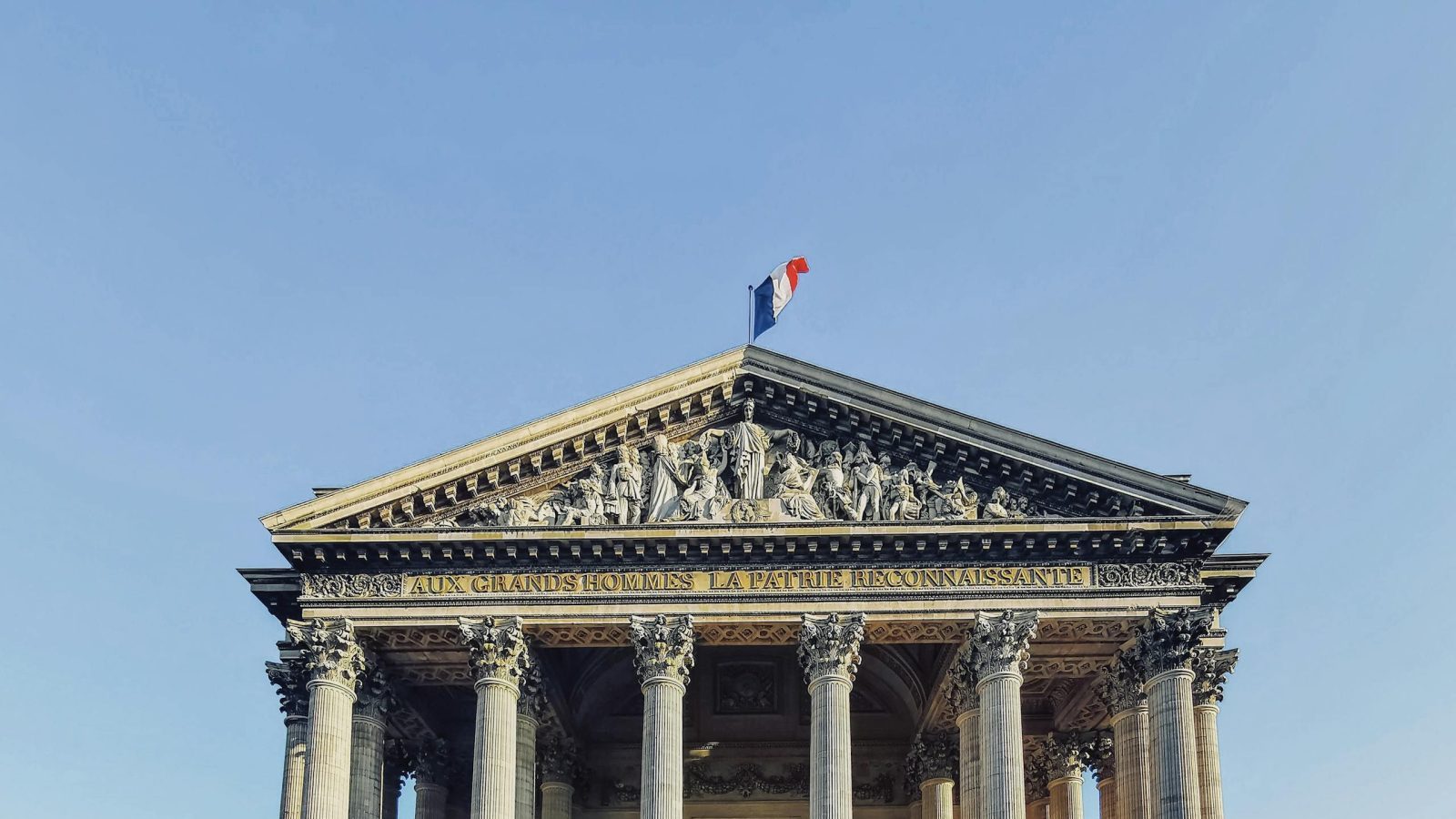 Front view of the Panthéon in Paris, featuring its iconic columns and French flag under a clear blue sky.