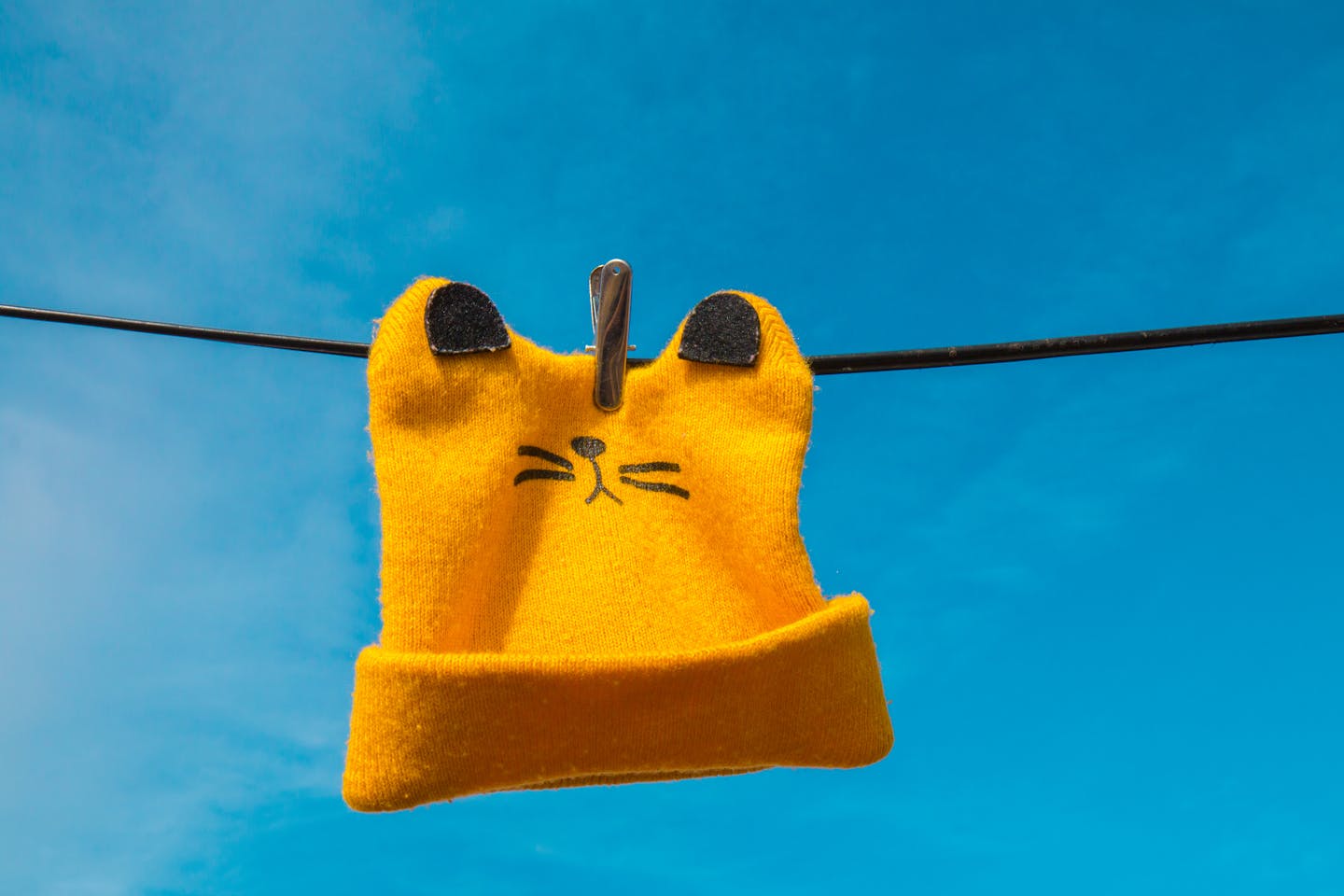 Bright yellow beanie with ears and whiskers design hanging on a clothesline against a blue sky.