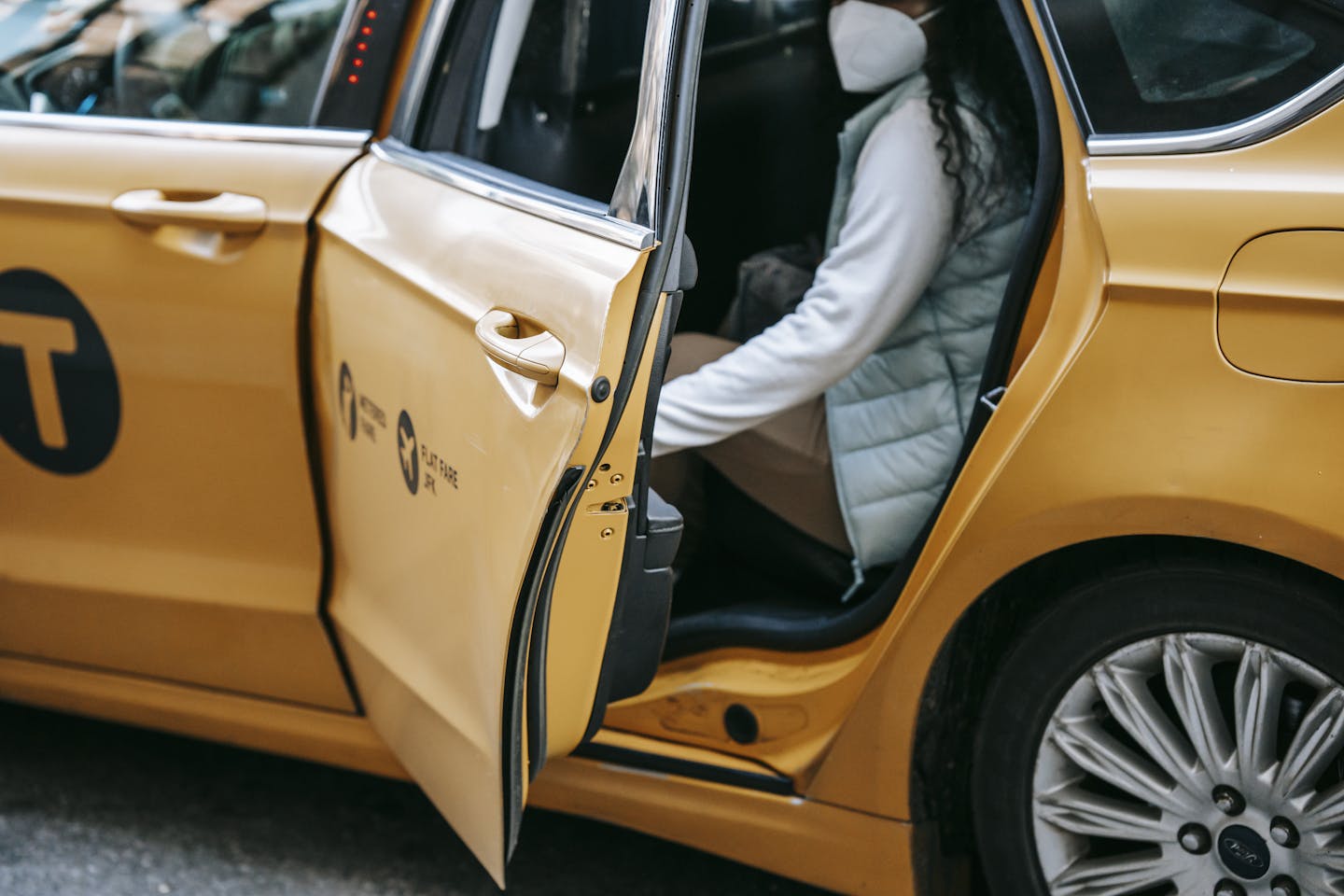 Side view of crop anonymous female in casual clothes and face mask sitting in taxi with opened door on street
