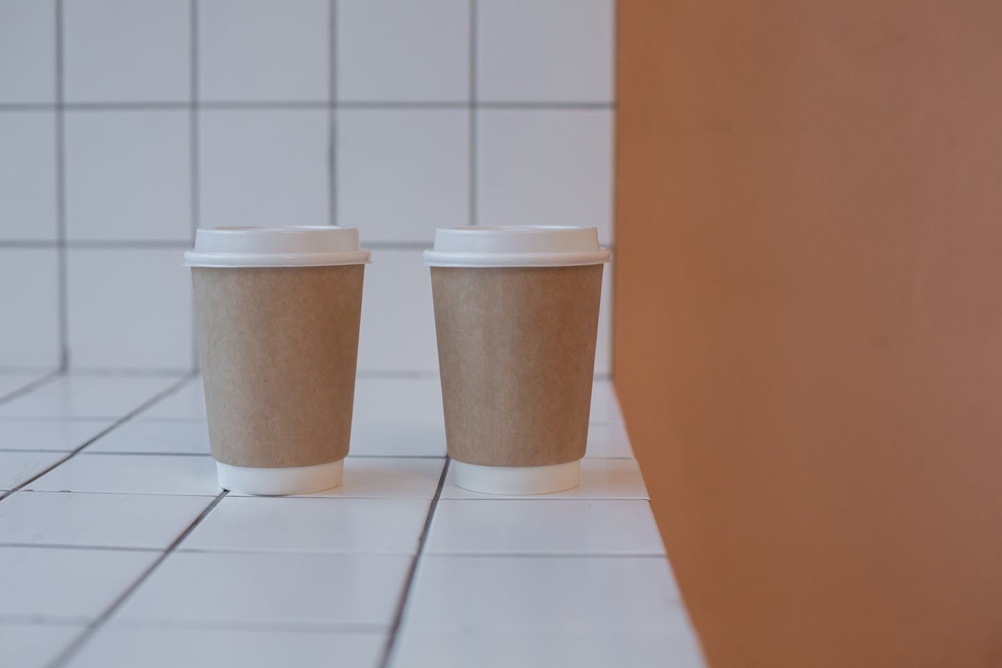 Two disposable coffee cups on a white tiled surface against an orange wall, minimalist style.