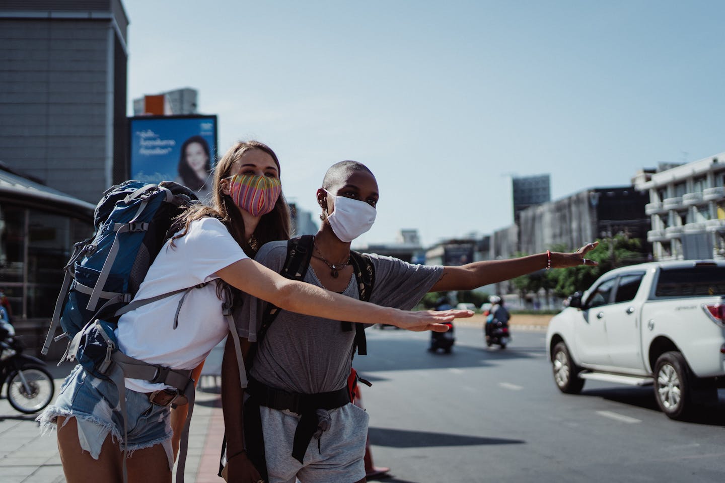 Two women with backpacks hitchhiking on a sunny day in a city wearing face masks.