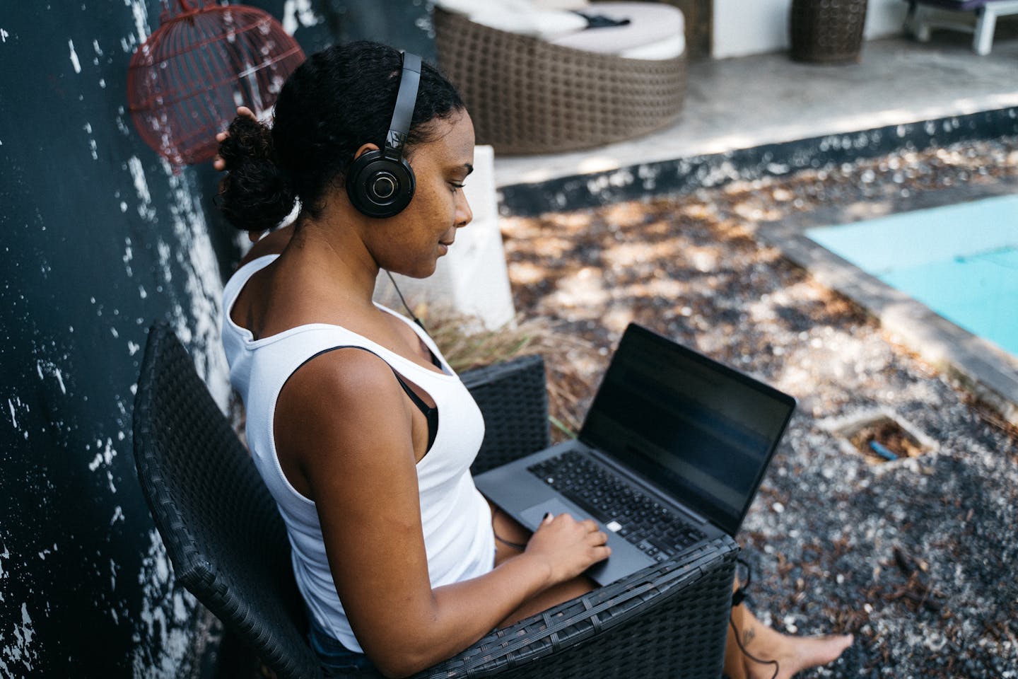Woman sitting by the pool with headphones, working on a laptop in a relaxed outdoor setting.