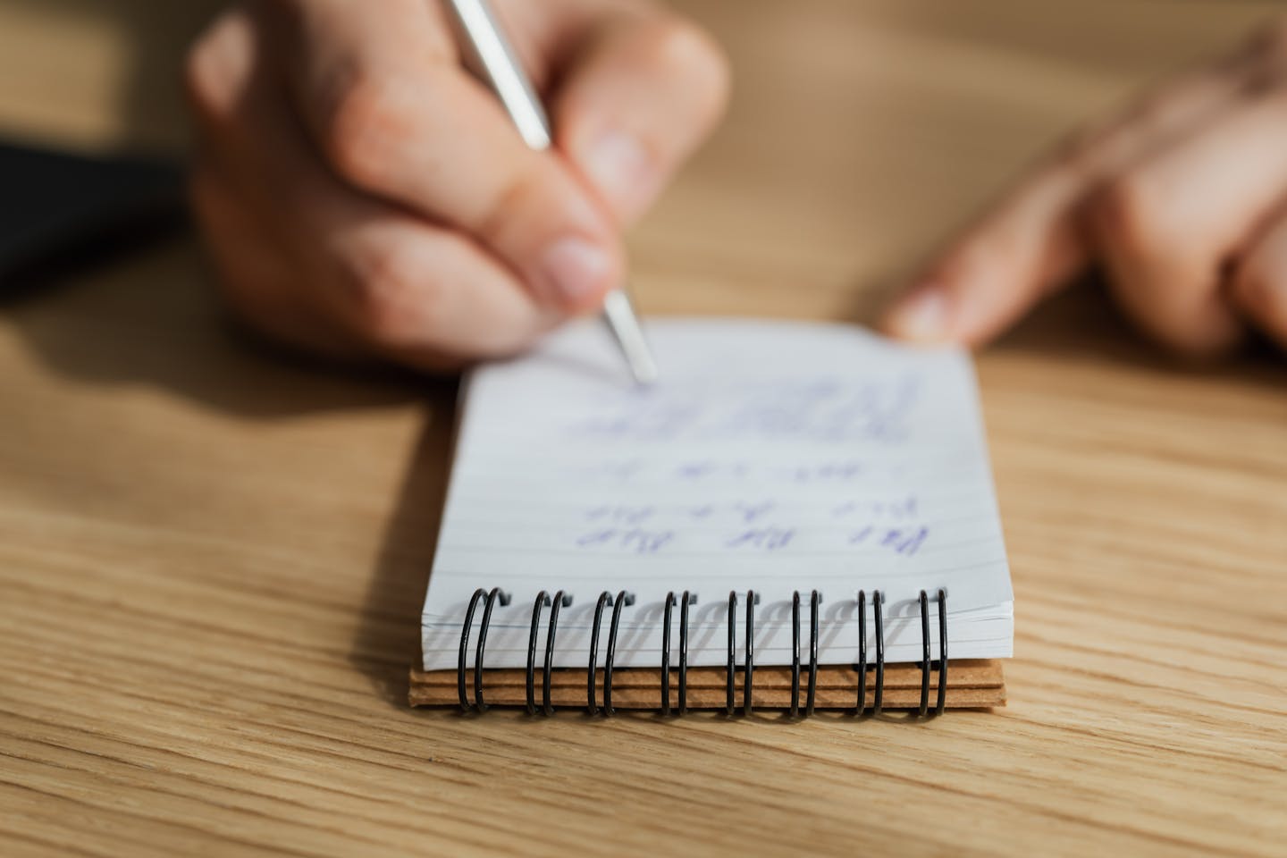 A focused moment capturing a hand writing notes in a spiral notebook on a wooden table.