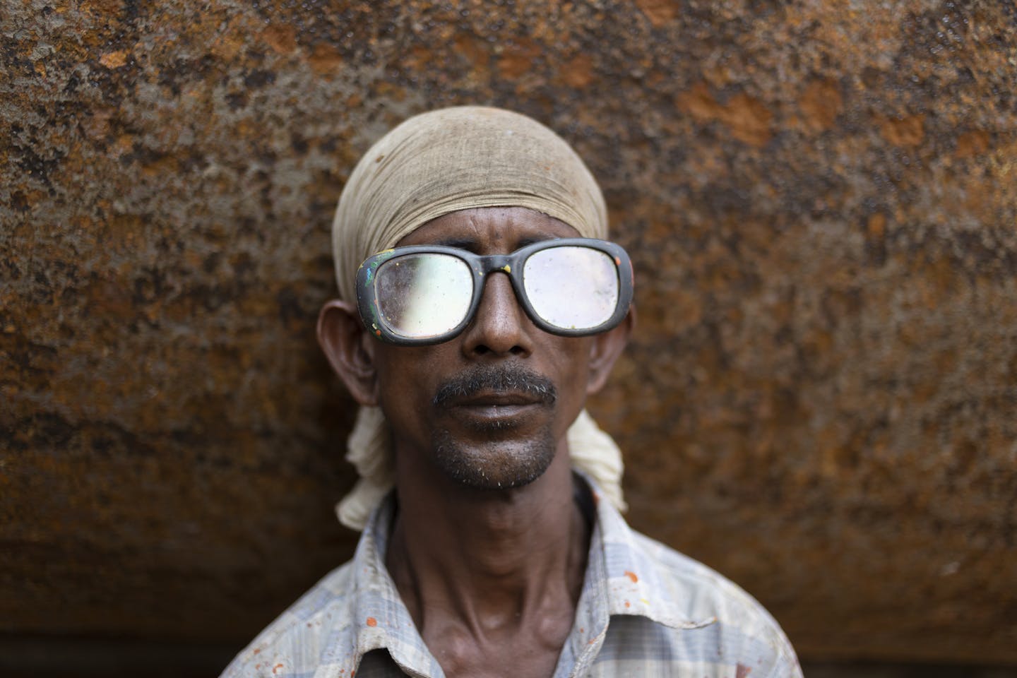 A man with a headscarf and sunglasses stands against a rusty background.
