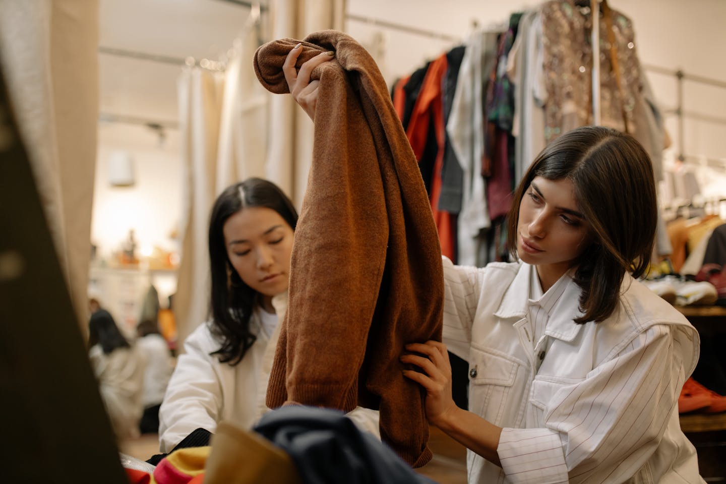 Two women browsing vintage clothing in a thrift store setting, examining garments closely.