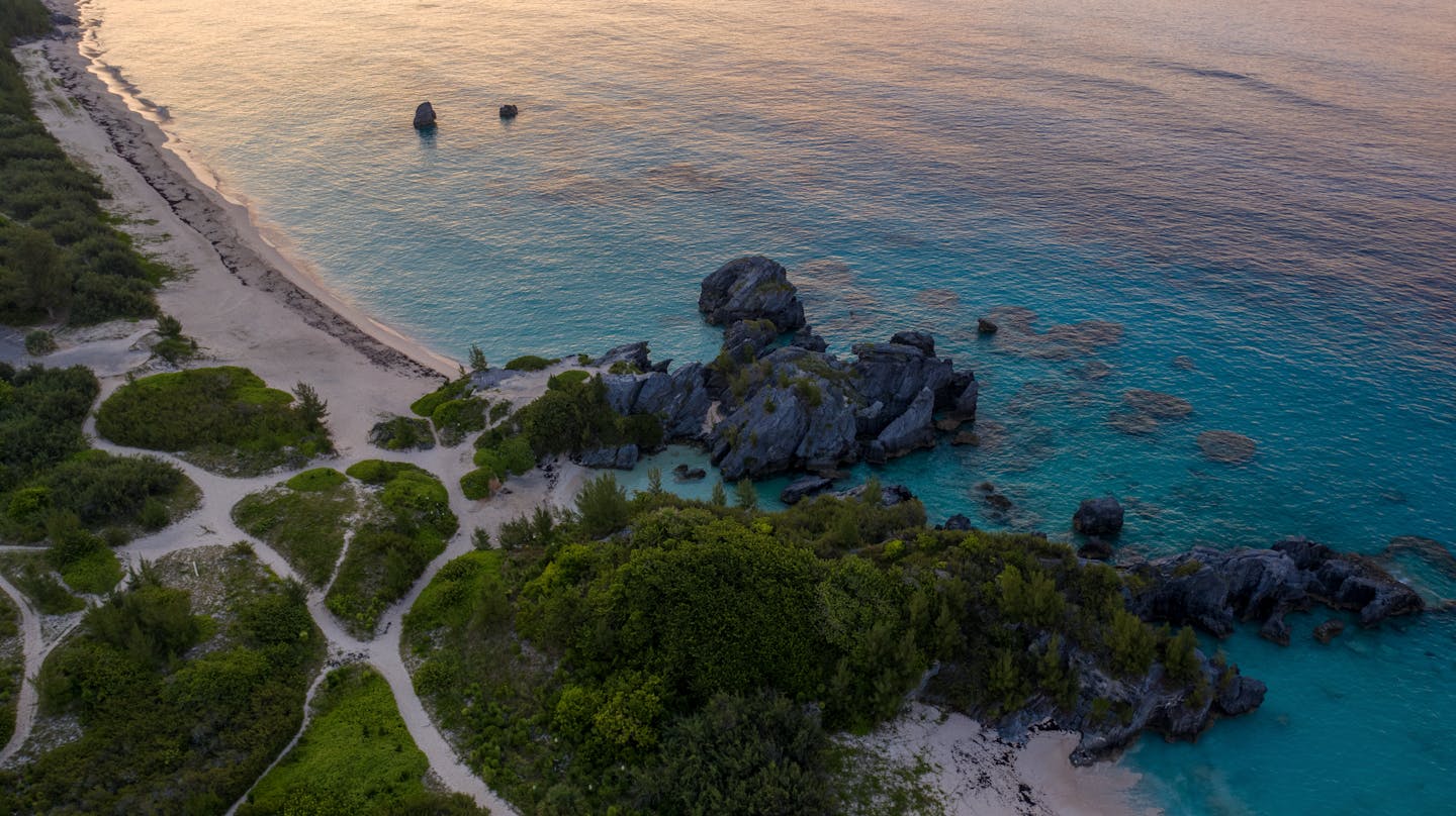 A breathtaking aerial view of Bermuda's coastline during sunset, showcasing clear blue waters and rocky formations.