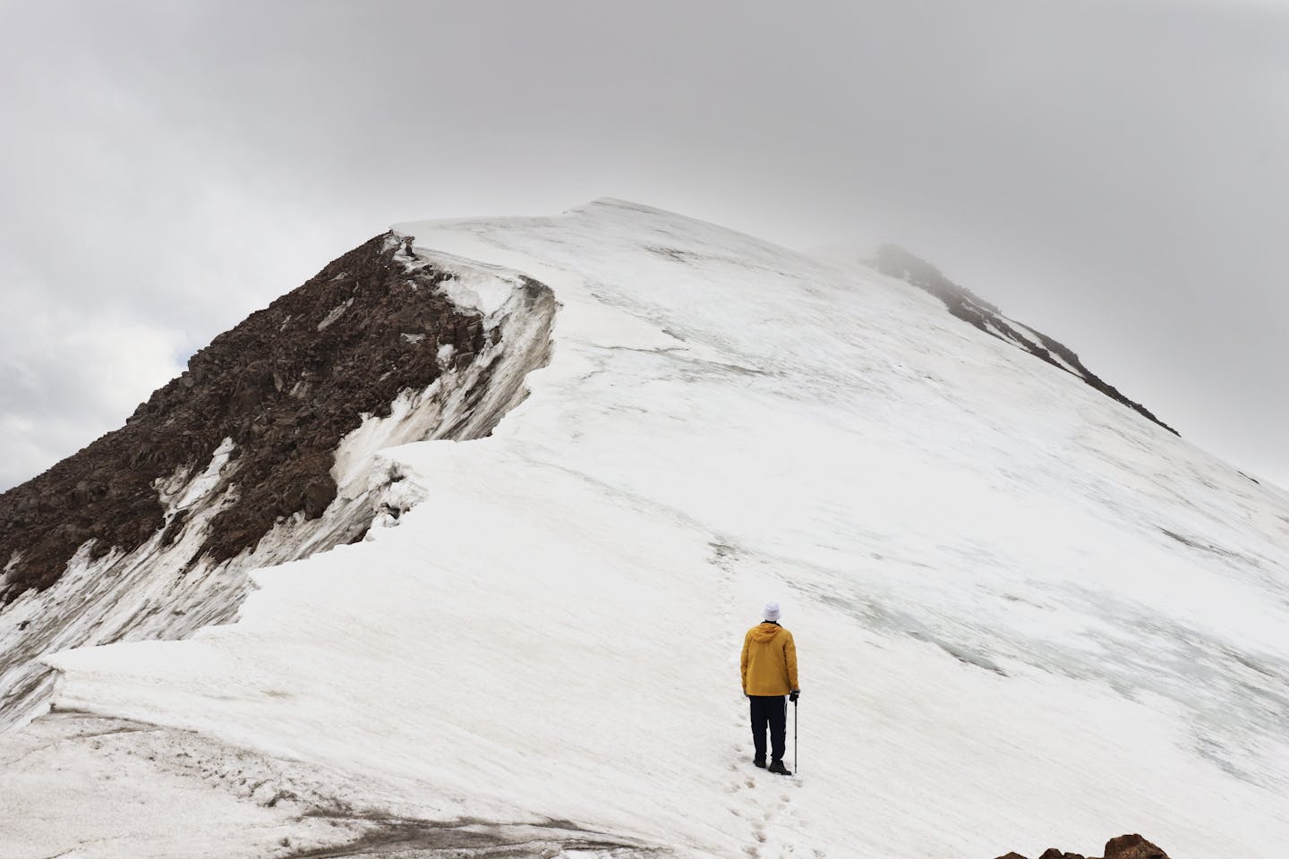 A lone climber in winter gear ascends a snowy mountain peak under cloudy skies.