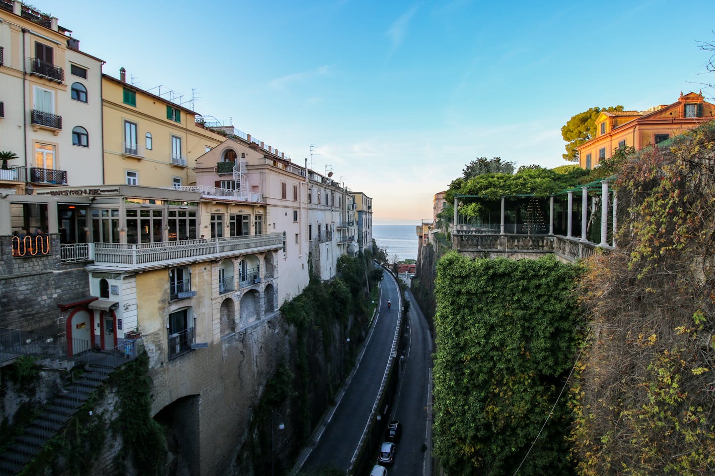 Charming cliffside buildings overlook a coastal road and the sea in Sorrento at sunset.