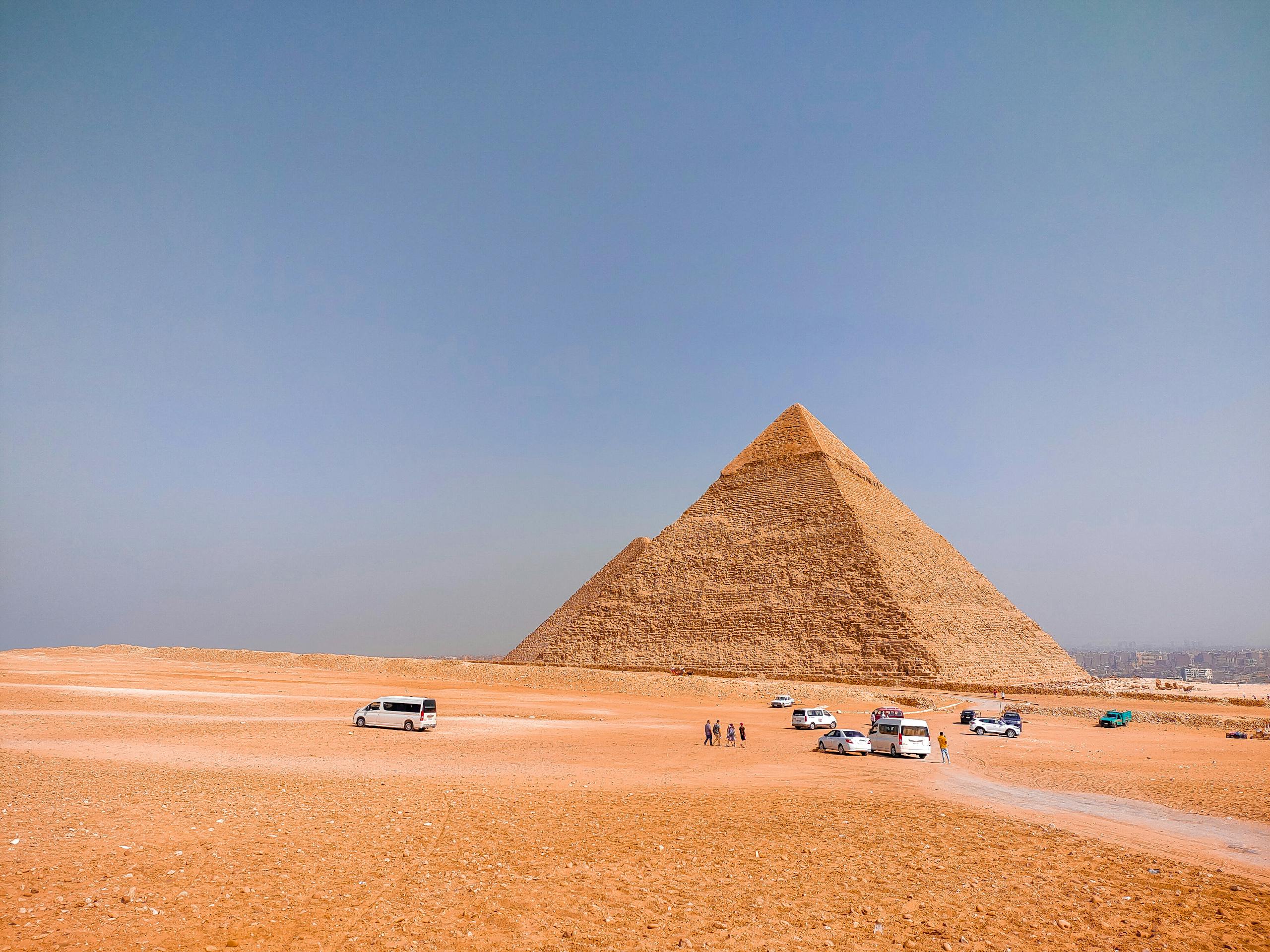 View of the Great Pyramid of Giza, surrounded by tourists and cars under a blue sky.