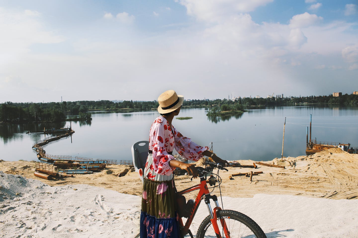 Woman in straw hat and floral dress biking beside a lake with a scenic view.