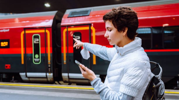 teenage girl in warm jacket, points to arriving train on station platform, holding mobile phone in hands
